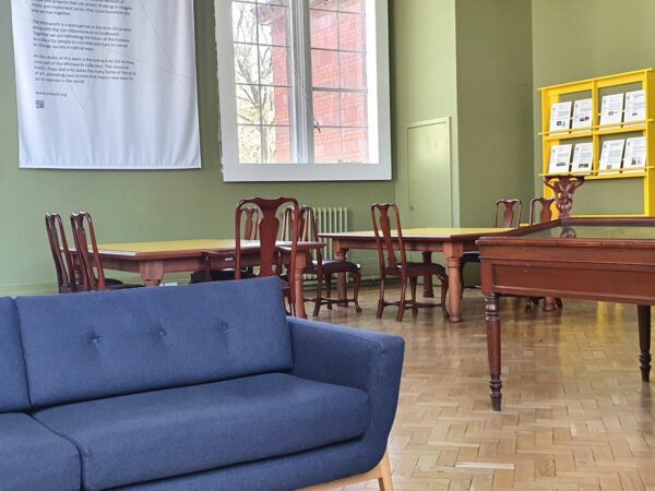 A room with light blue walls, a big window, several wooden tables and chairs, a blue couch in the forefront.