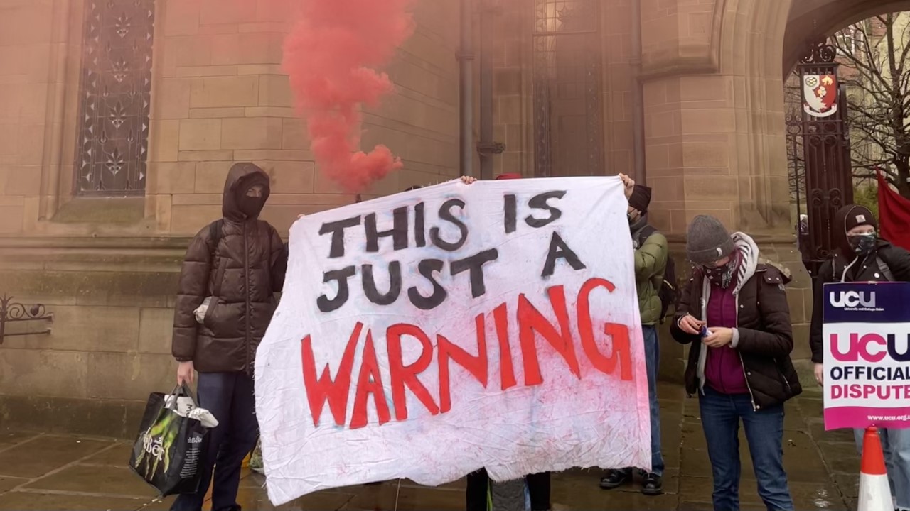 Occupiers from the John Owens Building stand in front of the Whitworth Arch holding a sign that says 'This is just a warning', and let off a flare with red smoke.