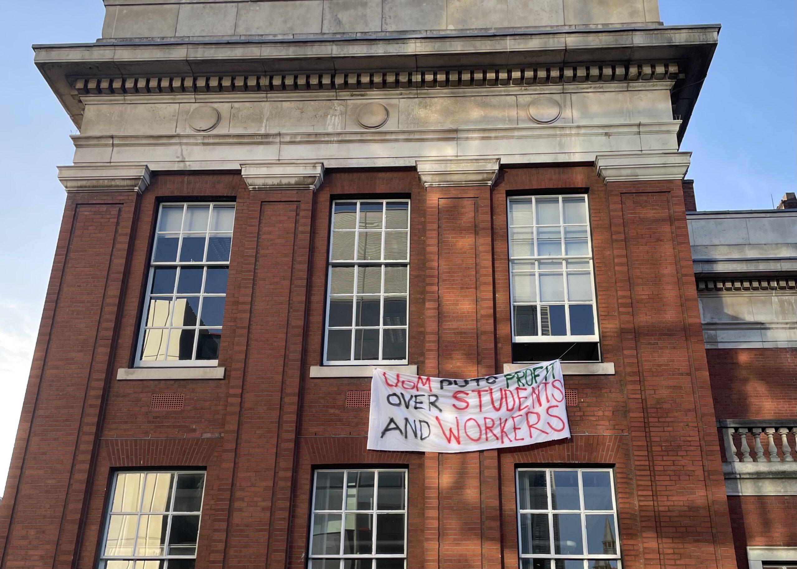 The Samuel Alexander Building with a banner hanging out of the window. The banner says "UoM puts profit over students and workers".