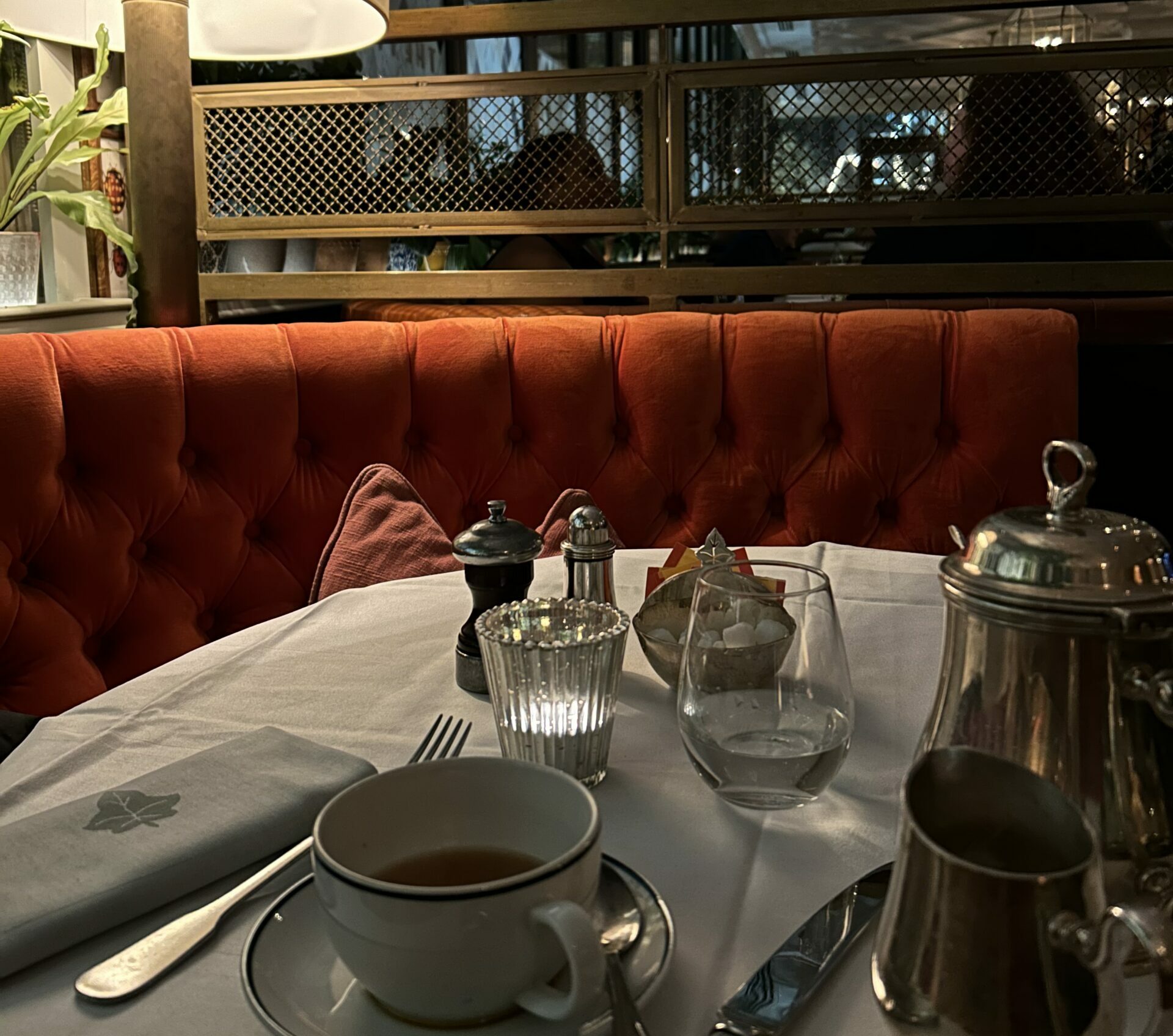 This image shows a silver pot of tea, a white ceramic teacup and saucer, upon a dining table in The Ivy restaurant. Around the table is a red cushioned booth and some house plants.