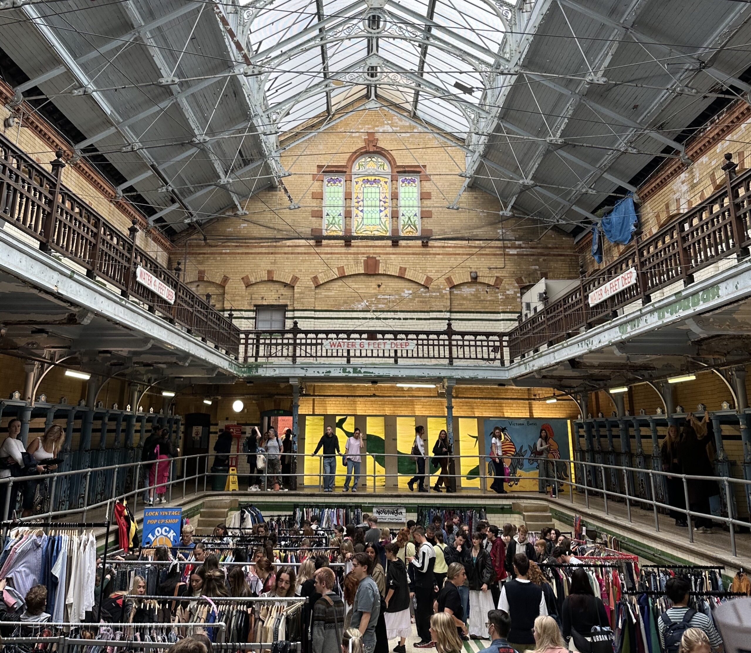 A tall interior of a building with a glass ceiling and stained glass windows. People browse racks full of clothes below.