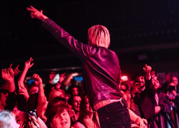 Oscar sings down at crowd level, standing on the barrier with his arm outstretched.