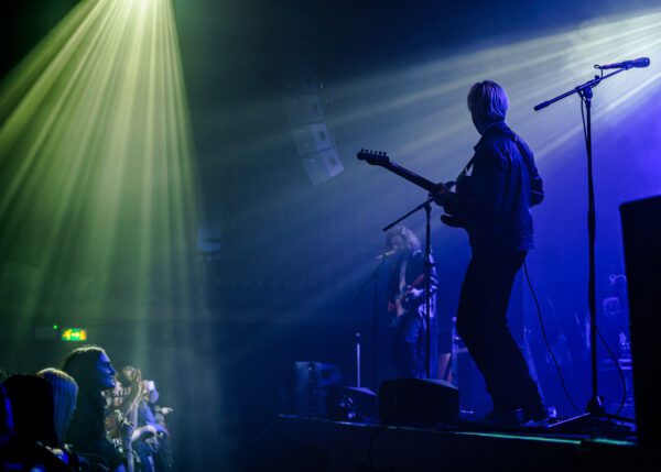 Sundara Karma on stage. Oscar plays guitar in silhouette. Ally plays guitar behind him, singing backing vocals.