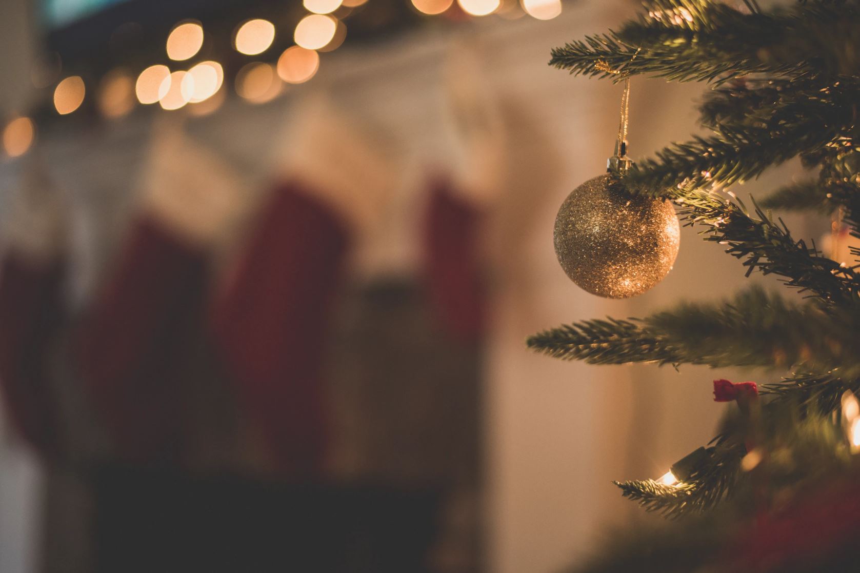 Close up of a gold bauble hanging on a Christmas tree with fairy lights and stockings in the background