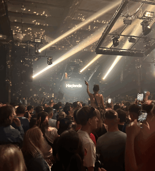 A large dancing crowd with bright lights. 'Hacienda' is on a screen at the front of the crowd. 