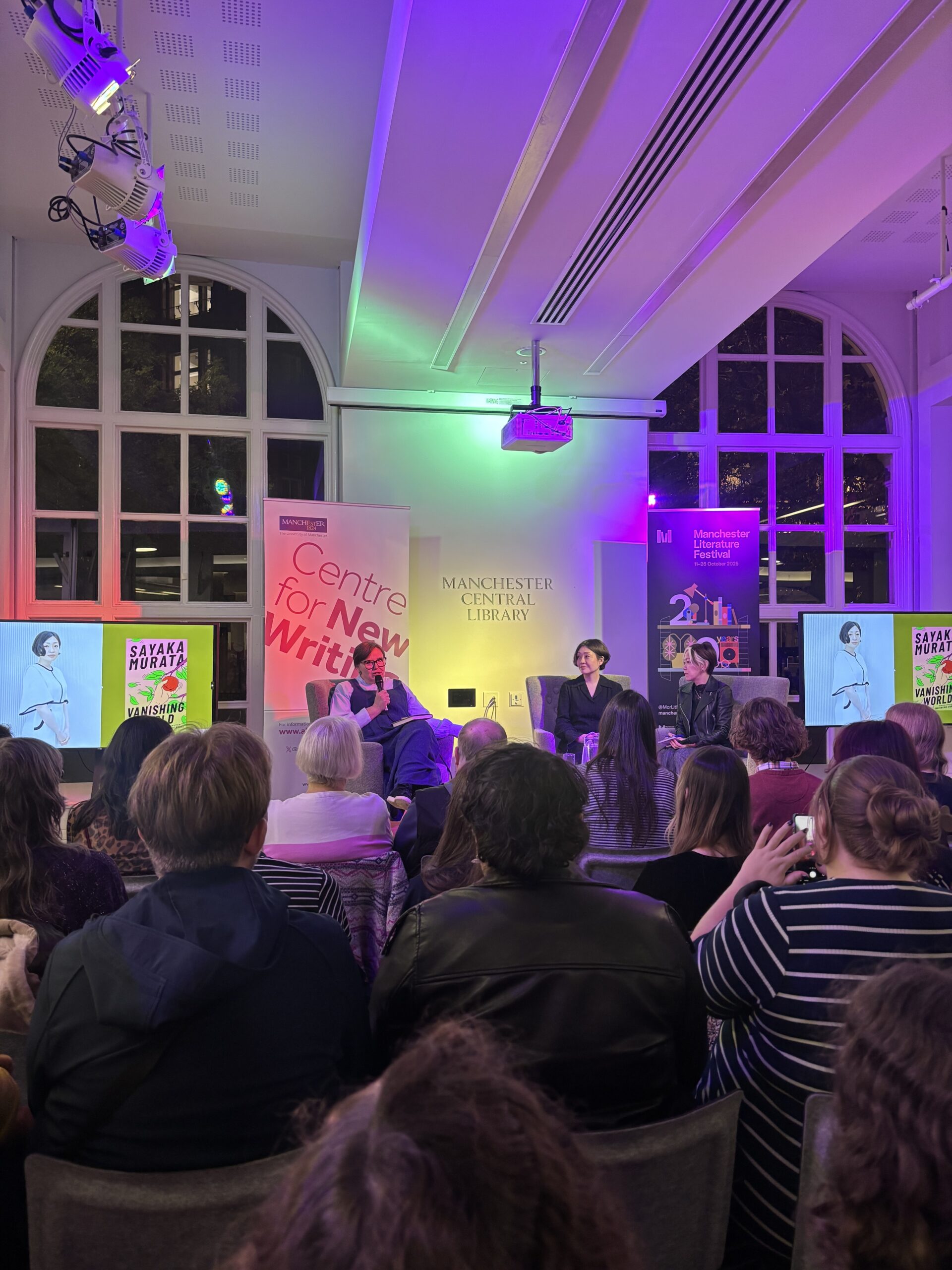 Three women - Sayaka Murata, her translator, and the interviewer - are on stage at Manchester Literature Festival. A crowd is seated in front of them, with screens to the side depicting Murata's book 'Vanishing World.'
