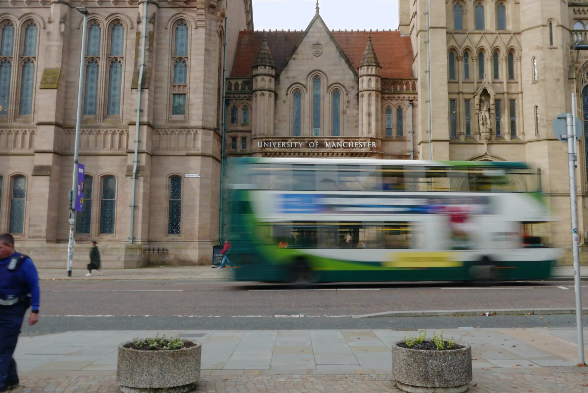 A yellow and green Bee Network bus with motion blur drives past UoM's Whitworth Building on Oxford Road, Manchester.